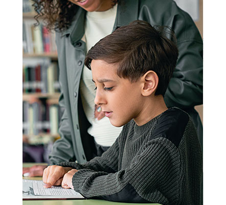 young boy focused on reading a book, with a teacher standing behind him